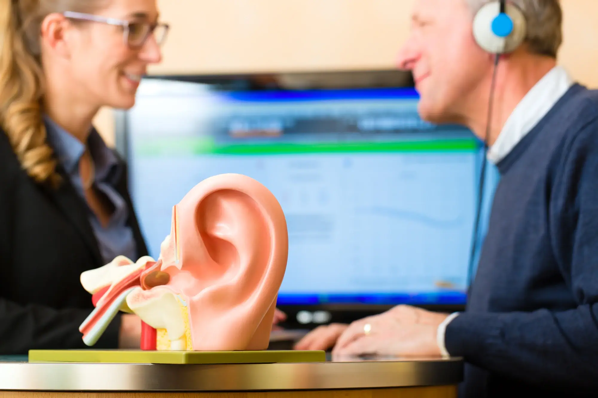 A human ear model with people doing a hearing test blurred in the background