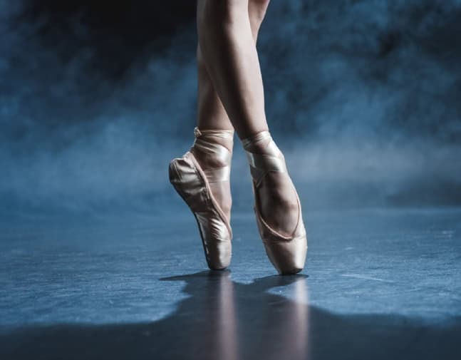 A cropped view of a ballet dancer in pointe shoes in a dark dance studio.