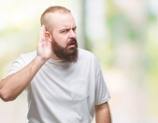 Man wearing over isolated background with hand over ear listening having trouble hearing with a frustrated look on his face.