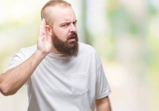 Man wearing over isolated background with hand over ear listening having trouble hearing with a frustrated look on his face.